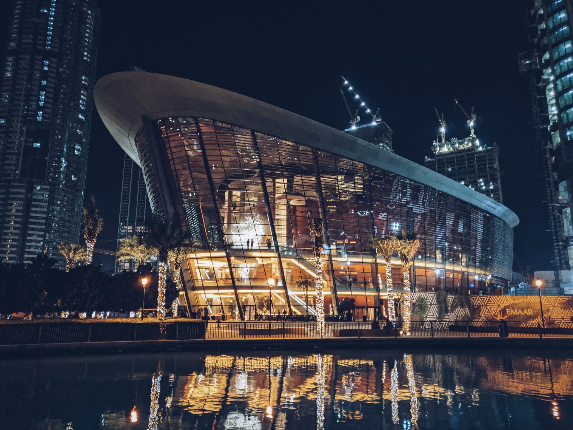 Modern, curved glass building at night with illuminated interiors and reflections in a nearby water body.